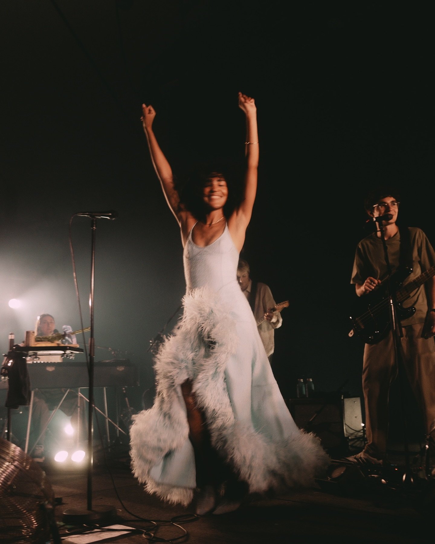Cantora em vestido azul dança animadamente no palco durante show com músicos ao fundo. Atmosfera vibrante.