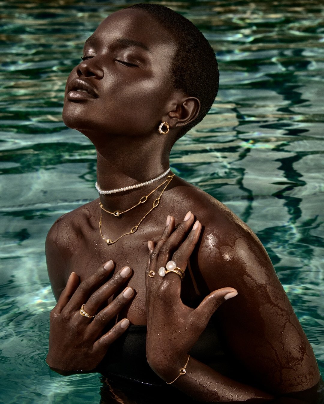 Mulher com joias douradas e collar de pérolas, em frente a uma piscina, em pose elegante. Pandora