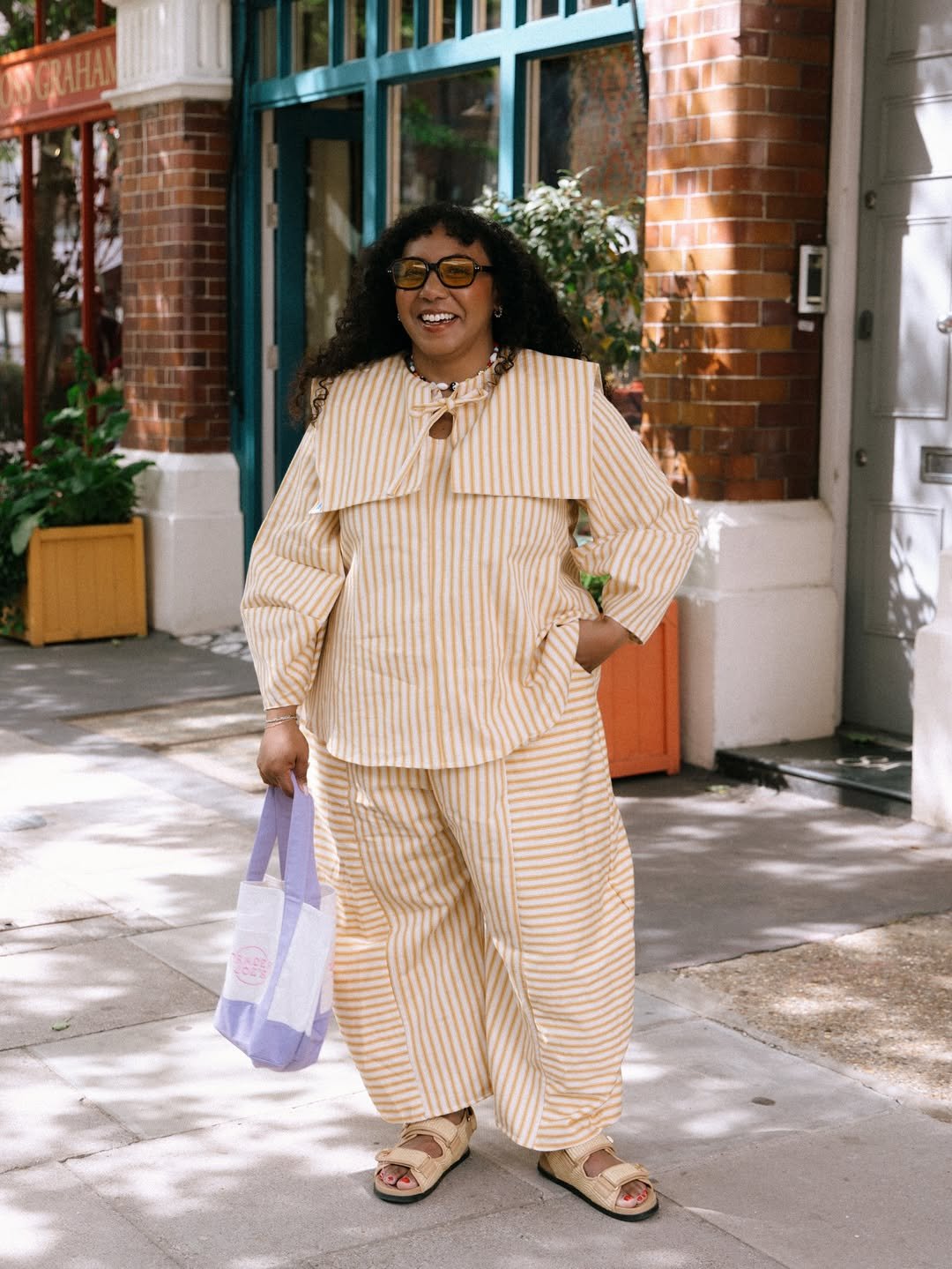 Pessoa sorridente vestindo roupa listrada amarela, segurando bolsa. Usa papete confortável, aclamada por seu estilo casual.