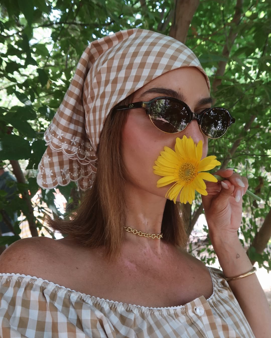 Mulher com lenço xadrez e óculos escuros segura flor amarela em dia ensolarado, trazendo os melhores perfumes árabes femininos