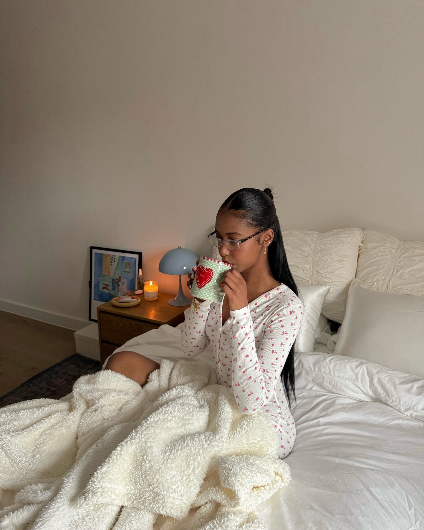 Pessoa em pijamas confortáveis no quarto, segurando caneca, cercada por cobertor macio. Ambiente acolhedor e relaxante.