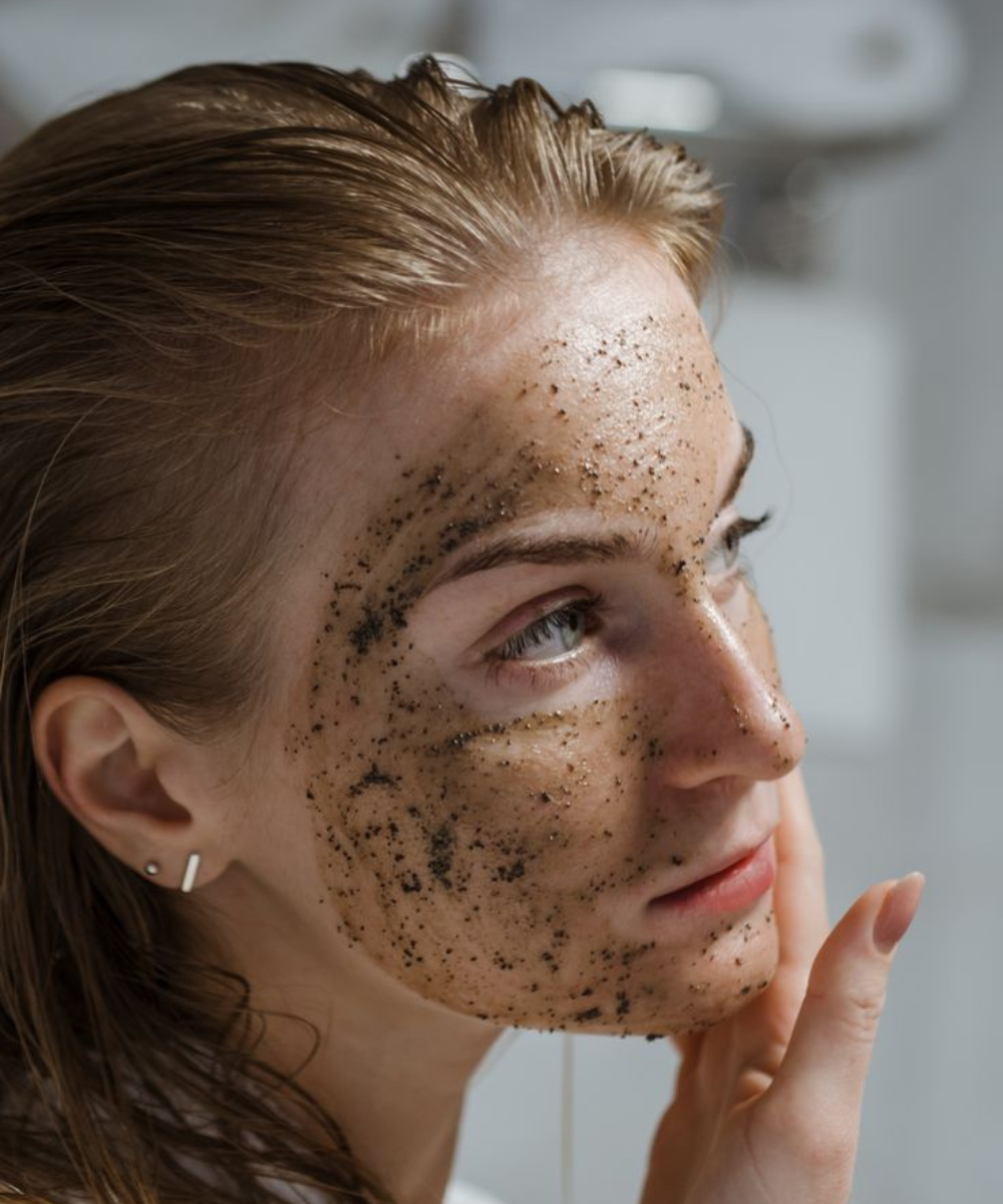 Mulher ensinando como esfoliar o rosto com máscara facial granulada, mão tocando a bochecha.