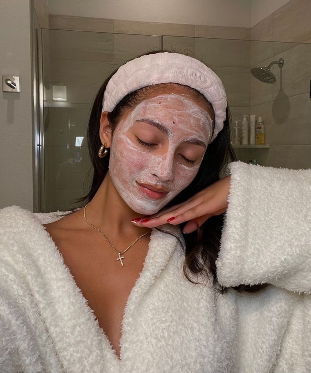 Pessoa com máscara facial descansando em um banheiro, usando roupão branco e faixa de cabelo.