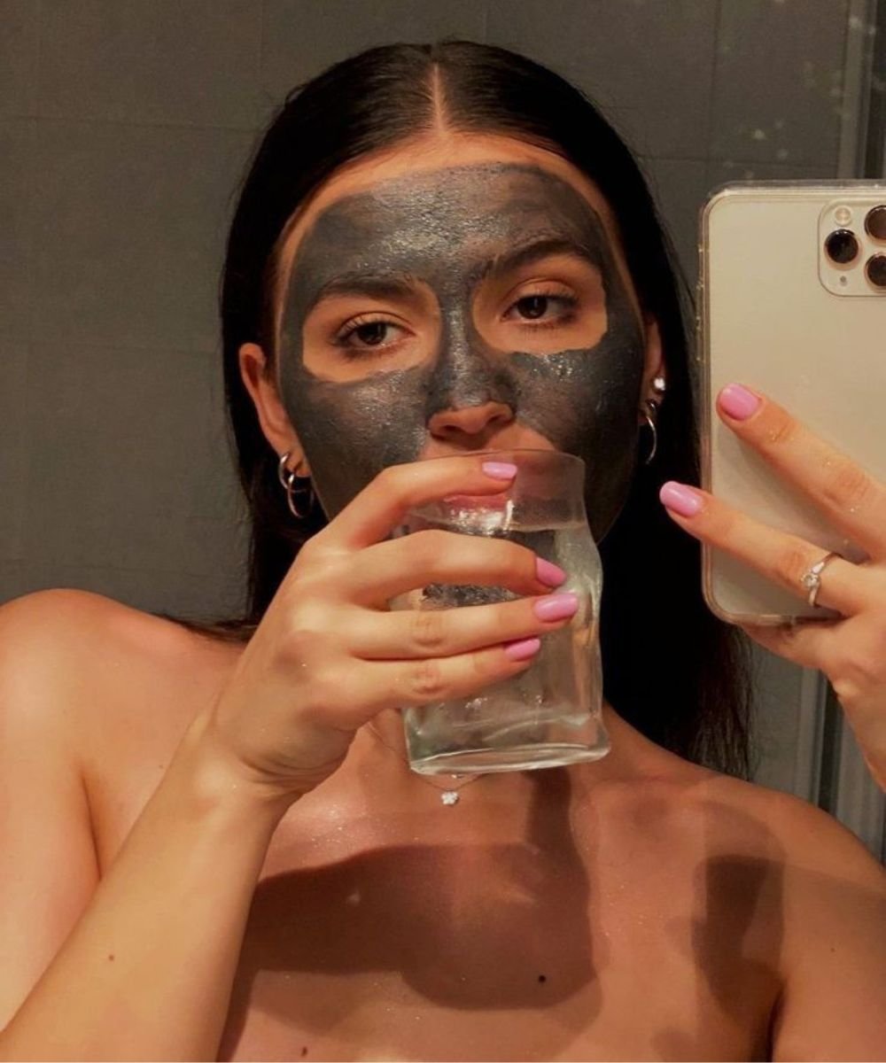 Mulher com máscara facial preta, tirando selfie enquanto segura um copo de água.