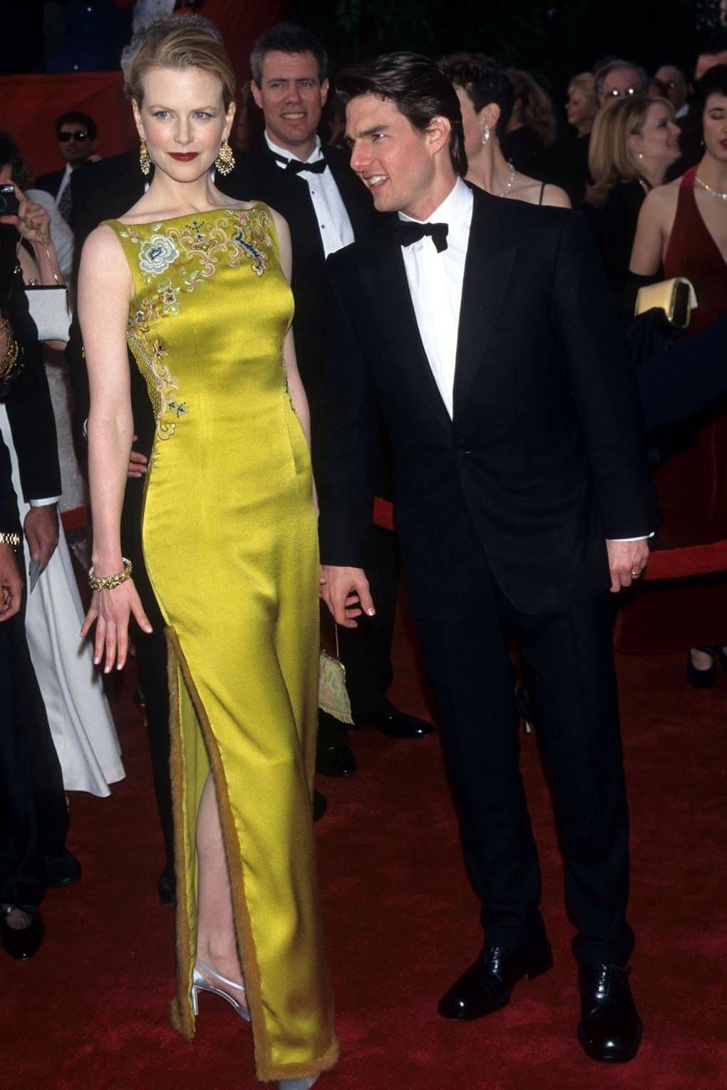 Casal elegante em um red carpet, ela com vestido amarelo e ele de smoking, cercados por outros convidados.
