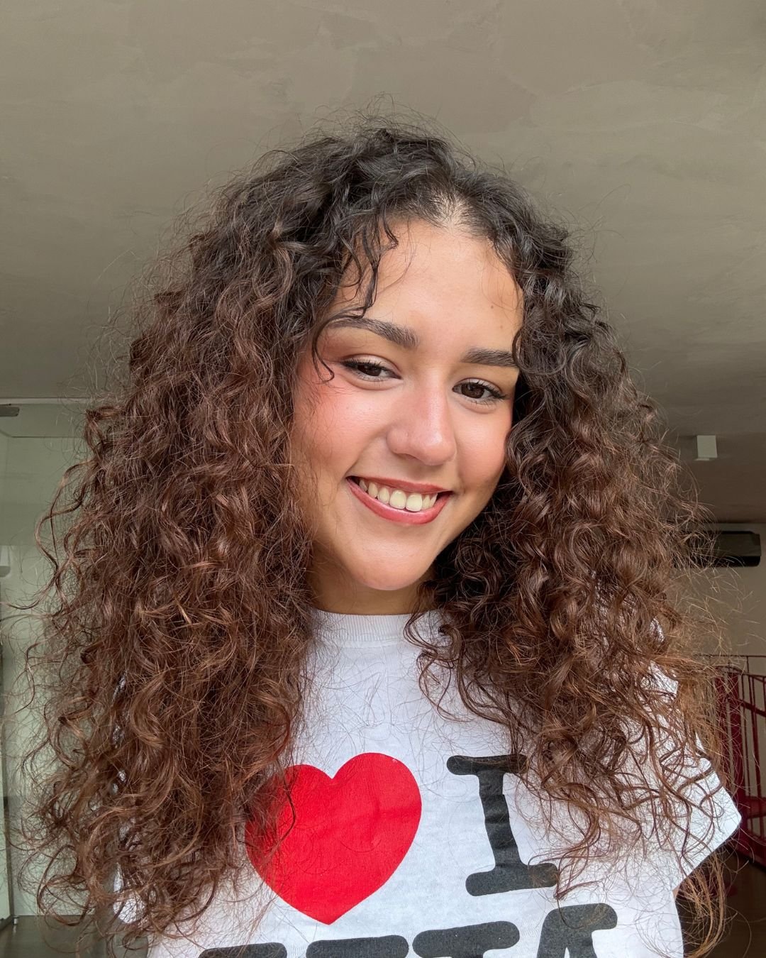 Pessoa sorrindo com cabelos cacheados volumosos, usando camiseta branca. O cabelo parece bem tratado com leave-in multibenefícios.