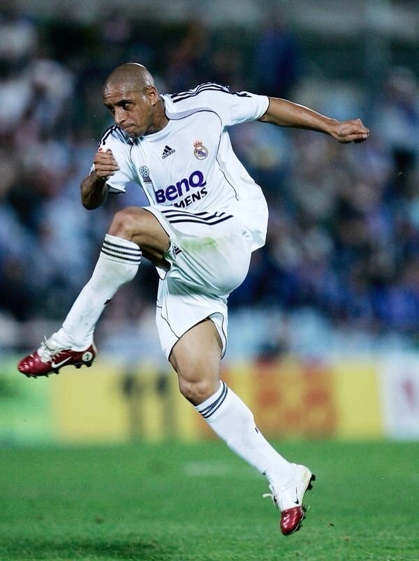 Jogador de futebol chutando a bola em campo, usando uniforme branco durante partida noturna.