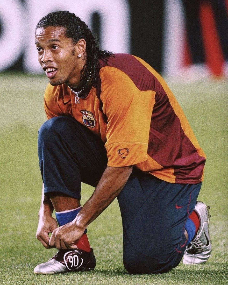 Jogador de futebol ajoelhado no campo amarrando chuteira com número 90, vestindo uniforme laranja e bordô.