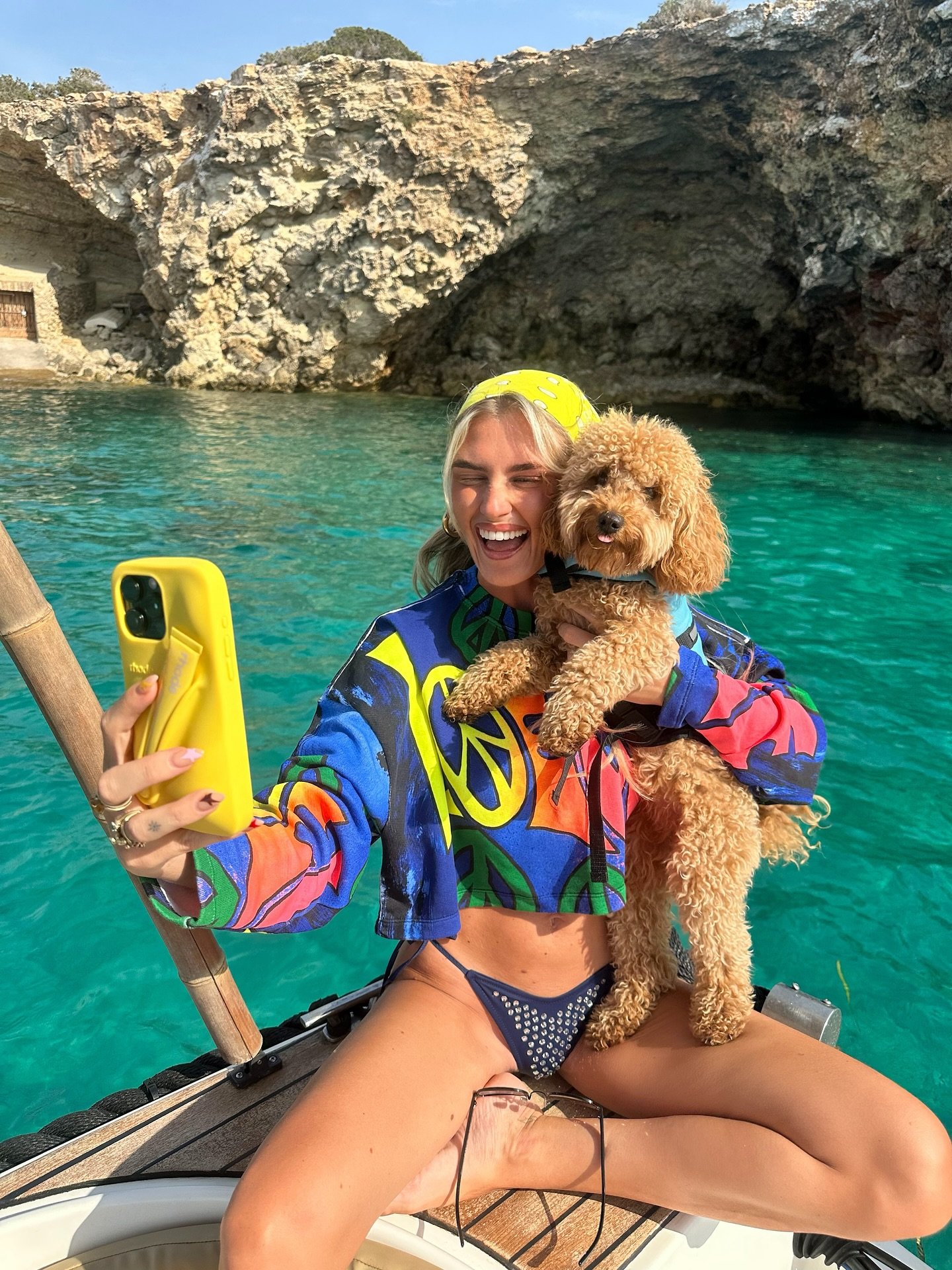 Pessoa em barco segurando cachorro com roupa colorida, cenário de mar e rochas ao fundo. Representando conteúdo sobre roupas para pets.