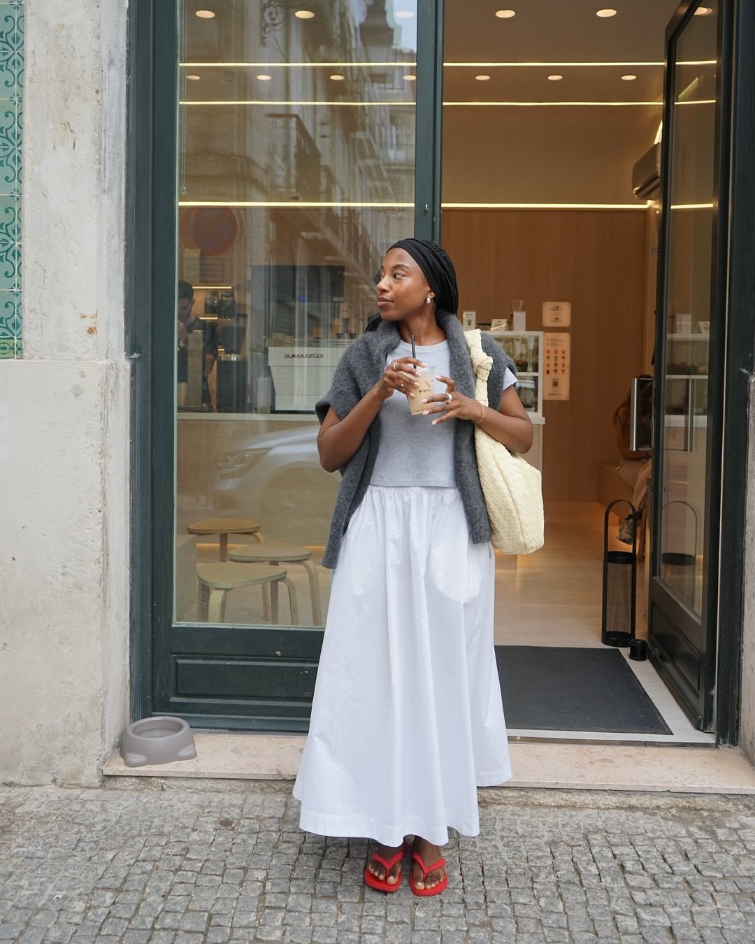 Mulher na rua usando saia longa e camiseta, segurando bebida em frente a uma loja.