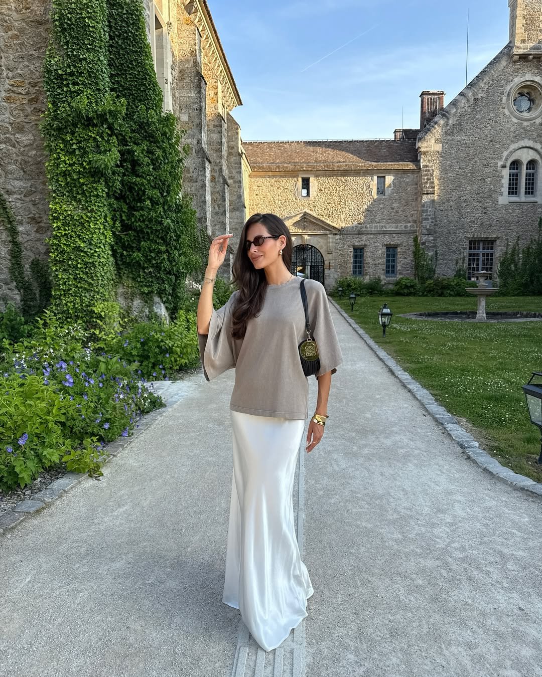Mulher usando saia com camiseta bege, caminhando em um jardim de prédio histórico com vegetação.