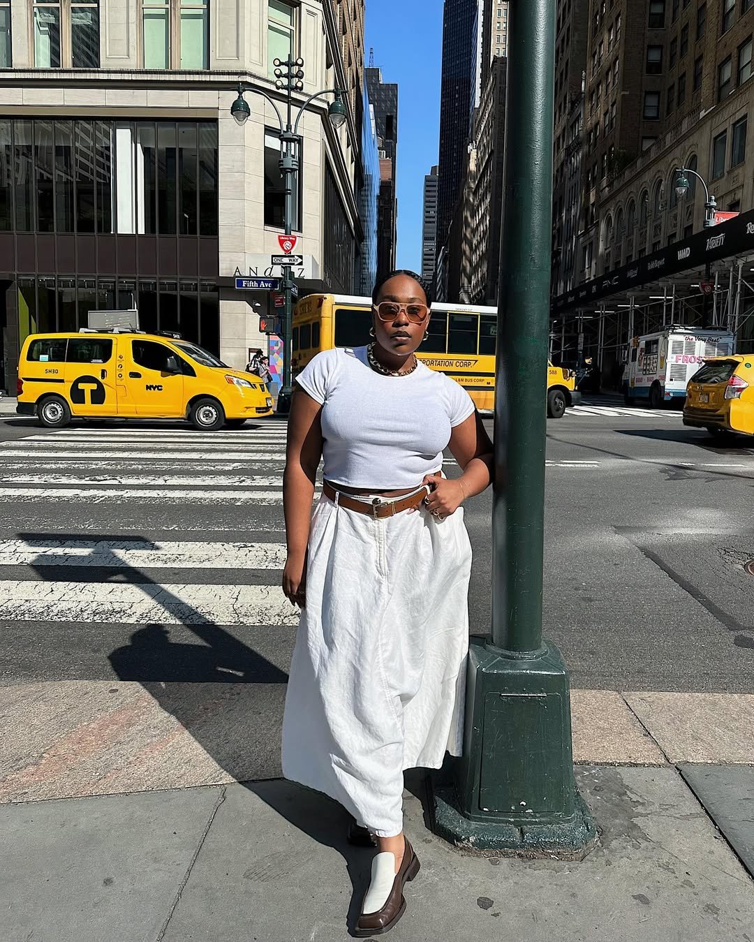Mulher na rua vestindo saia branca, camiseta e óculos escuros, cercada por táxis e prédios.