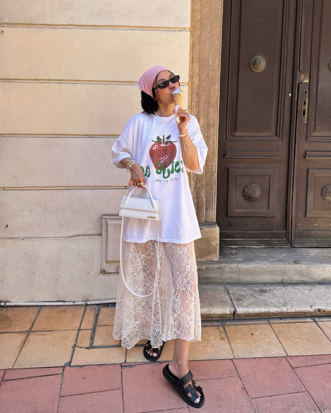 Mulher com saia de renda sobre calça, camiseta estampada e bolsa, saboreia sorvete na rua.