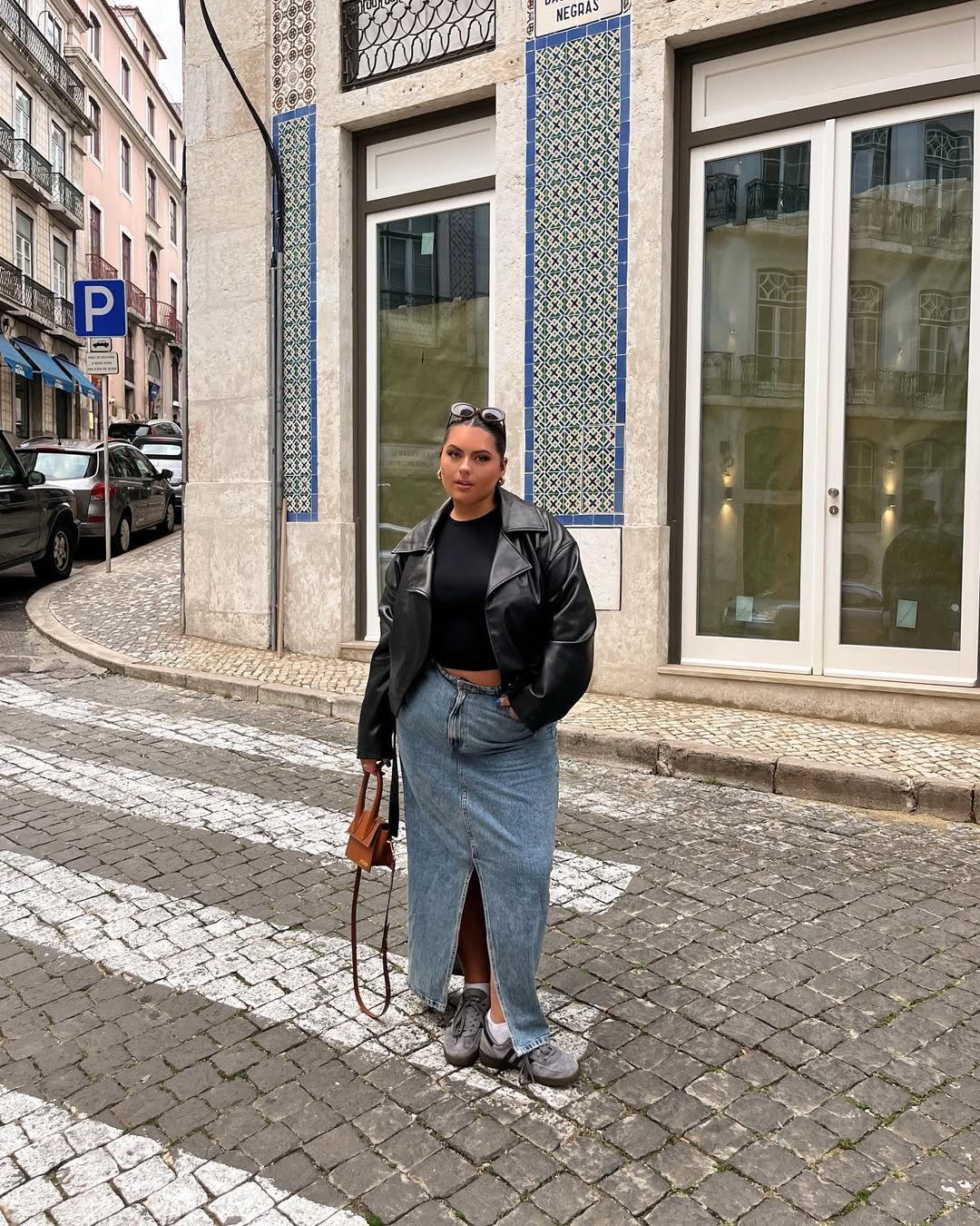 Mulher usa jaqueta preta e saia jeans longa com camiseta preta na rua, segurando bolsa marrom.