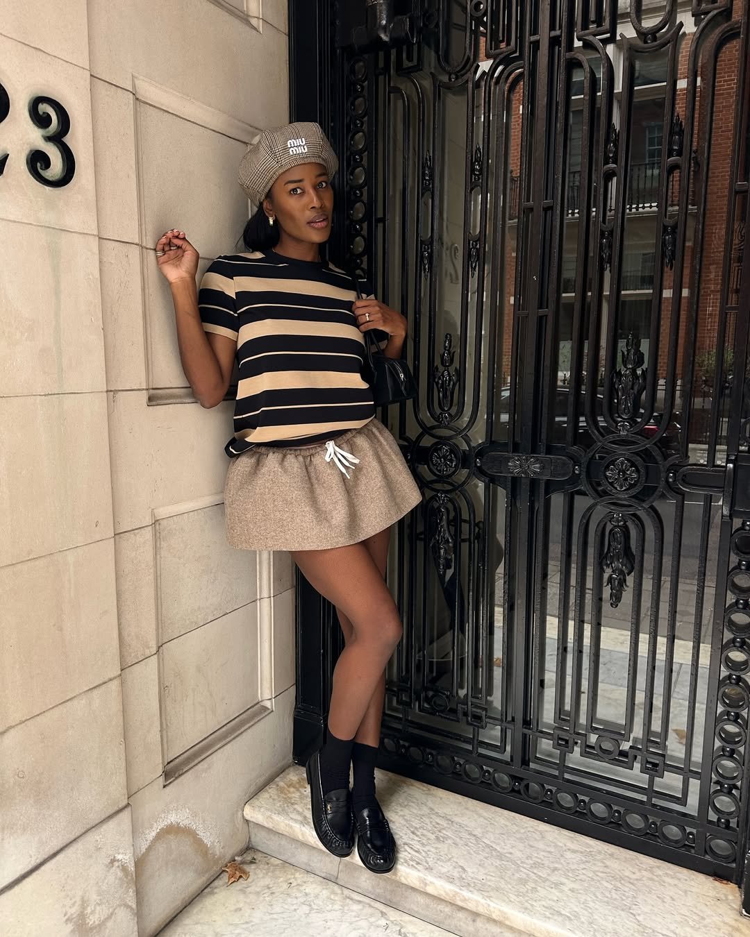 Mulher em pé, vestindo saia curta bege e camiseta listrada, encostada em porta de ferro ornamentada.