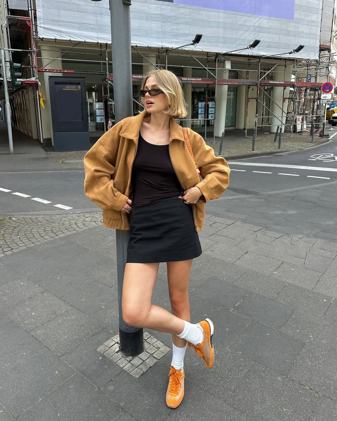 Pessoa loira com casaco caramelo e saia curta preta, óculos escuros e tênis laranja, em ambiente urbano.