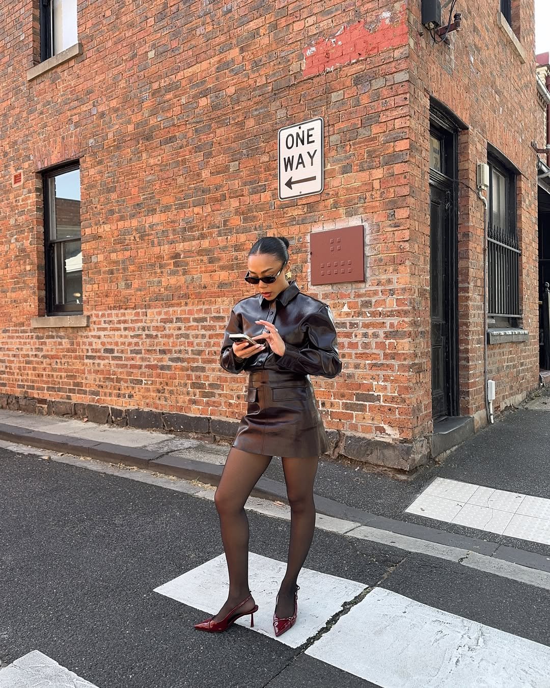 Pessoa em uma esquina urbana, usando casaco e saia curta marrom, consulta o celular. Há uma placa de 