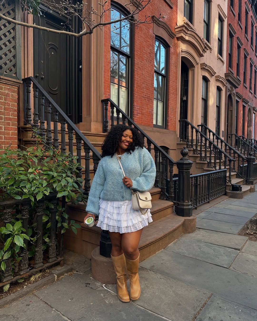 Mulher sorrindo na calçada em frente a prédio de tijolos, usando saia curta, suéter azul e botas bege.