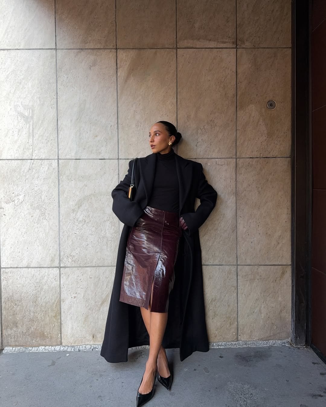 Pessoa vestida de sobretudo preto, saia elegante vinho e sapatos de salto, encostada em parede de pedra.
