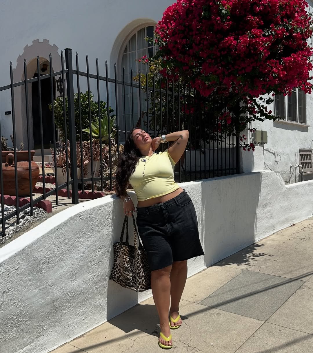Pessoa em pé, usando chinelos e roupa casual, posando ao lado de flores vermelhas e grade.