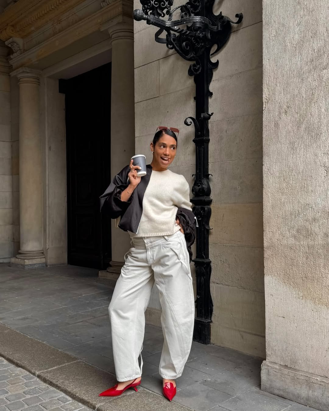 Pessoa com sapato vermelho de salto baixo, segura copo, veste suéter branco e calça larga, junto a parede de pedra.