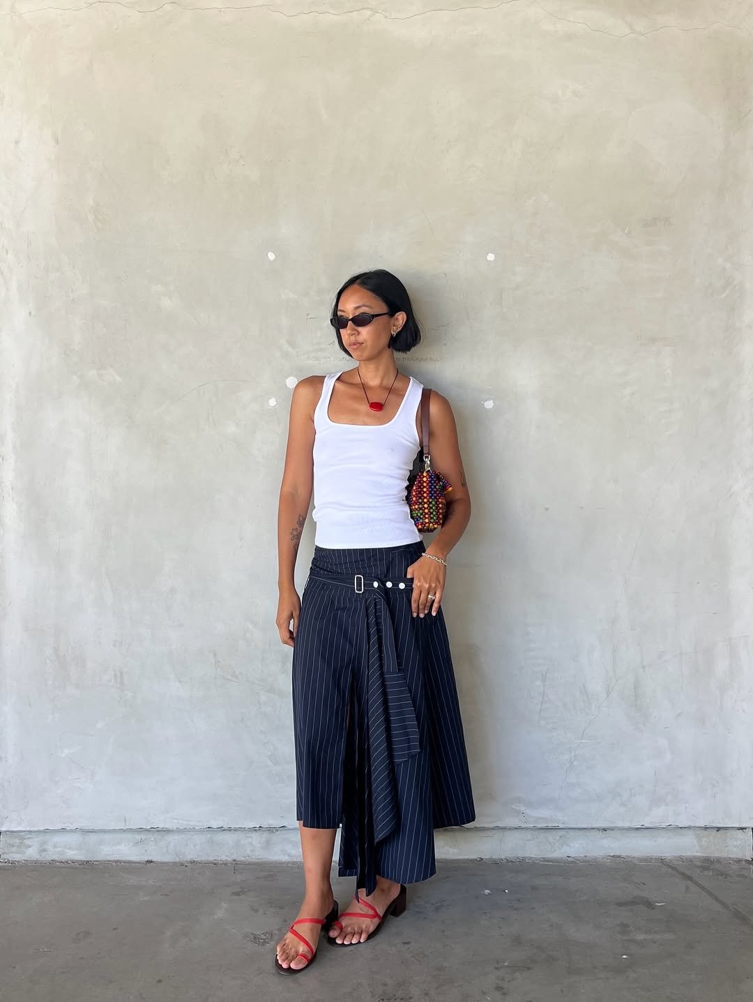 Pessoa com blusa branca, saia azul listrada, sapatos vermelhos e bolsa colorida, encostada na parede.