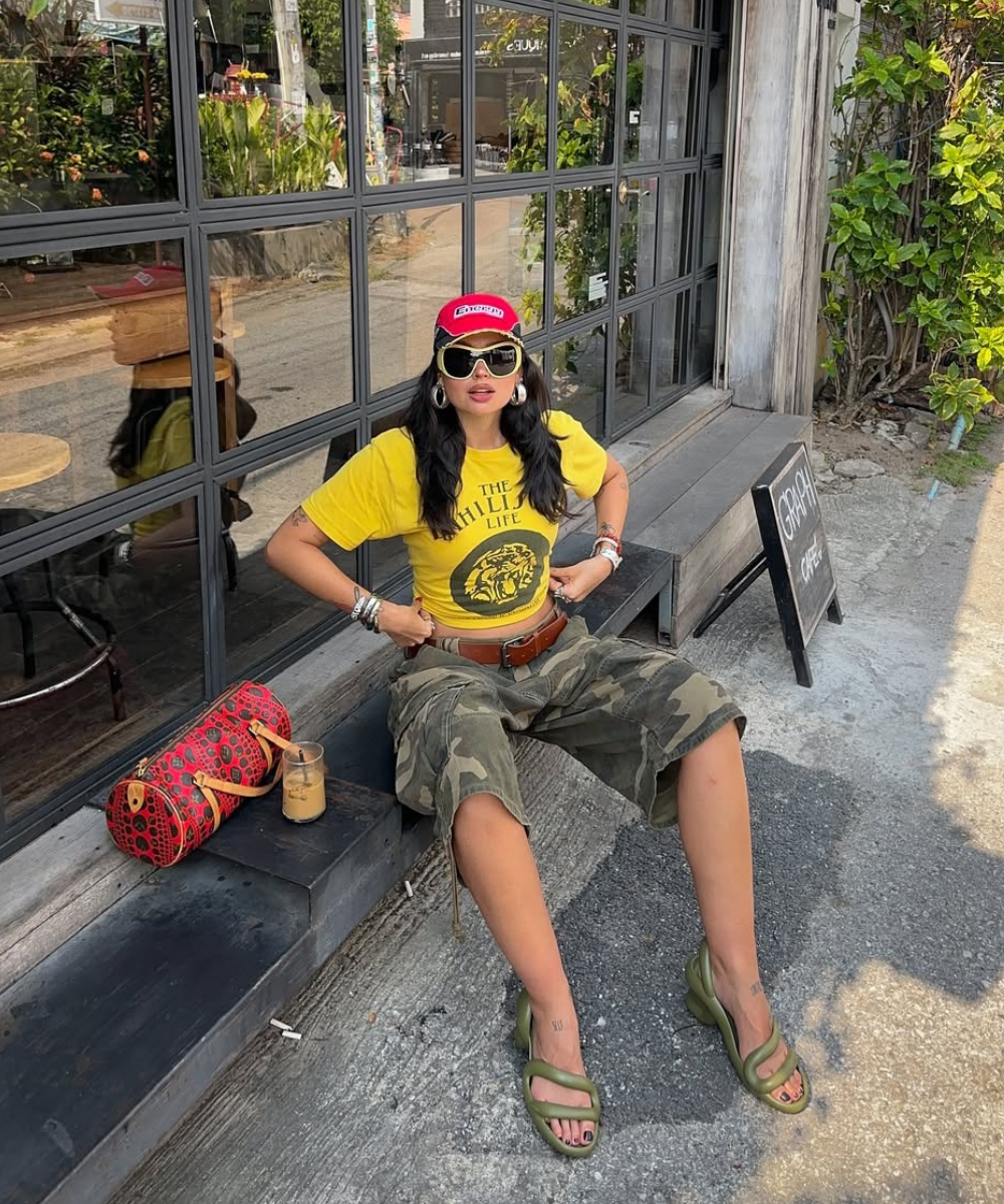 Mulher com camiseta amarela e bermuda camuflada sentada, usando sandálias estilosas verdes.
