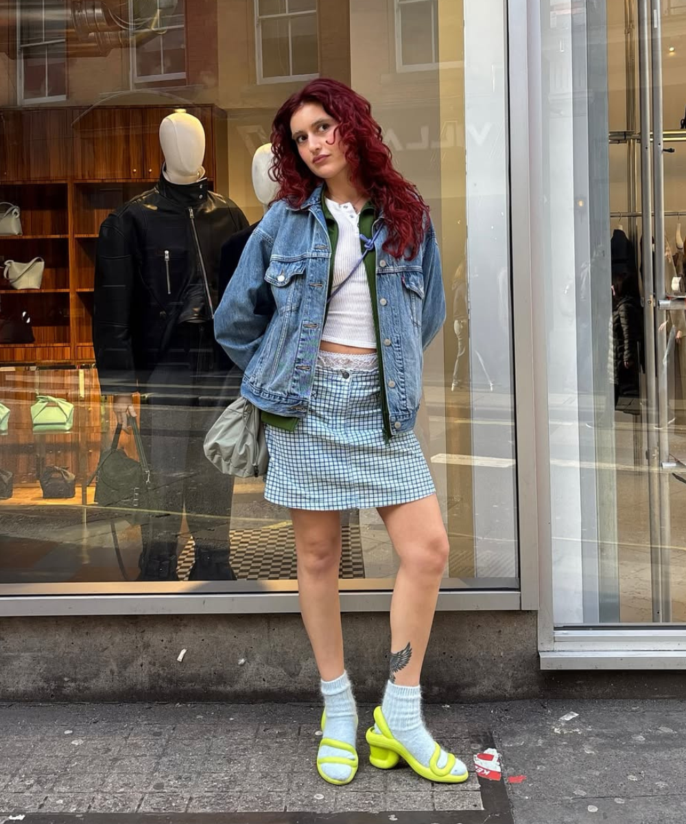 Pessoa com cabelo vermelho, jaqueta jeans, camiseta branca, saia xadrez e sandália colorida estilosa em frente a vitrine.