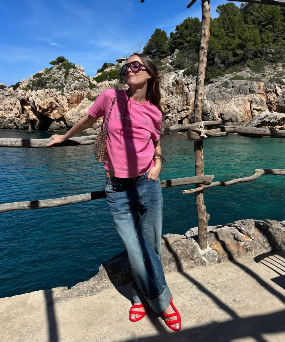 Mulher usando camiseta rosa, calça jeans e sandálias coloridas estilosas, em cenário de costa rochosa e mar.