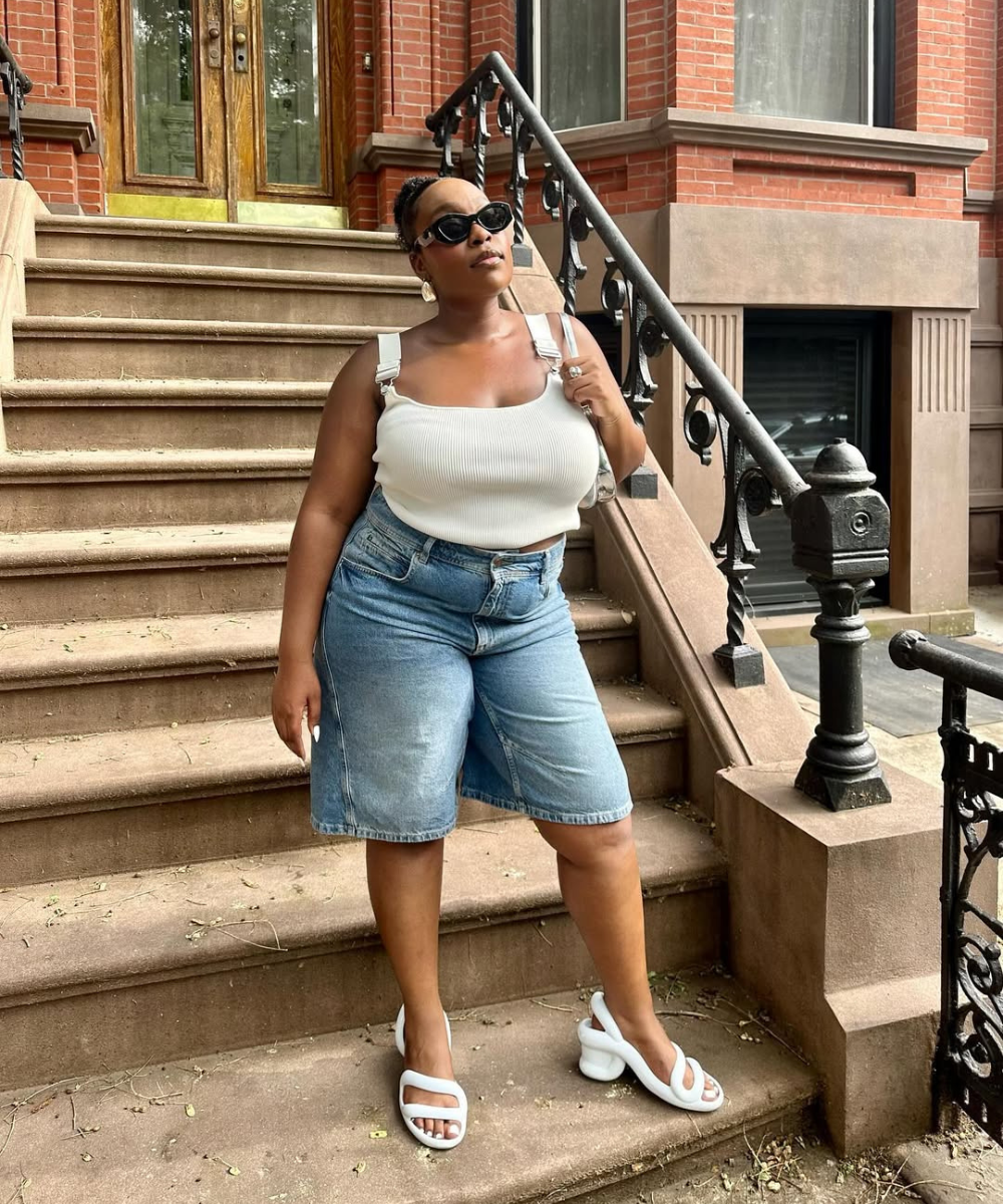 Pessoa em frente a prédio de tijolos, vestindo shorts jeans e blusa branca. Sandálias estilosas brancas.