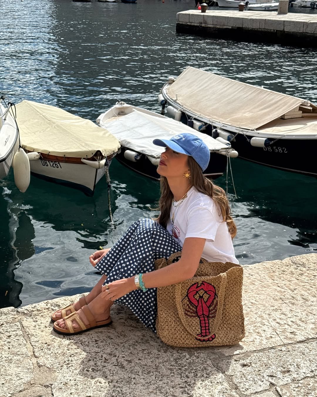 Sorrindo, mulher sentada em pier usa sandália de ráfia e segura bolsa de ráfia com desenho de lagosta, barcos ao fundo.