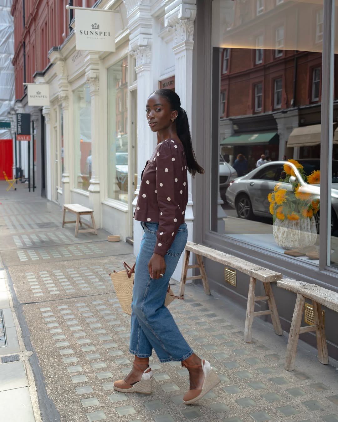 Mulher em rua elegante veste blusa marrom com bolinhas e sandália de ráfia, carregando bolsa de palha.