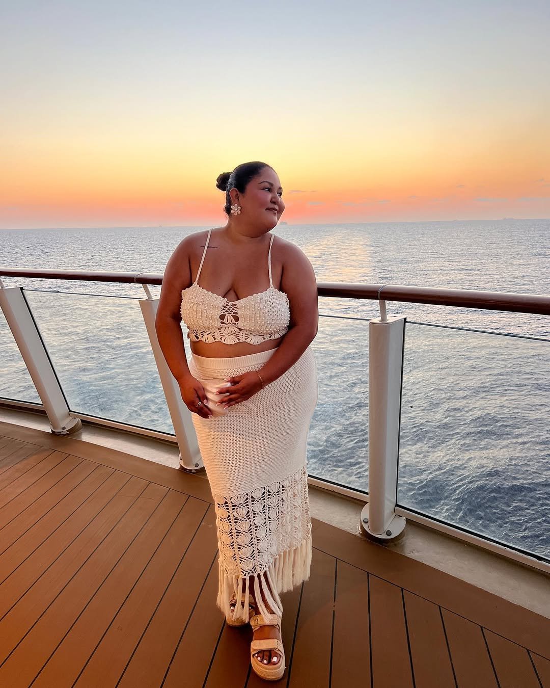 Mulher em cruzeiro ao pôr do sol, usando saia e top crochê com sandália de ráfia, diante do mar.