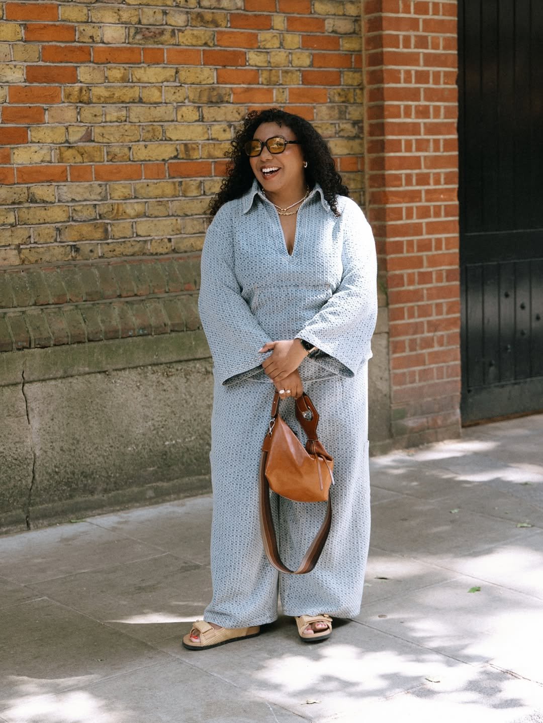Pessoa sorrindo com óculos escuros, vestindo conjunto azul e segurando bolsa marrom. Usa sandália de ráfia.