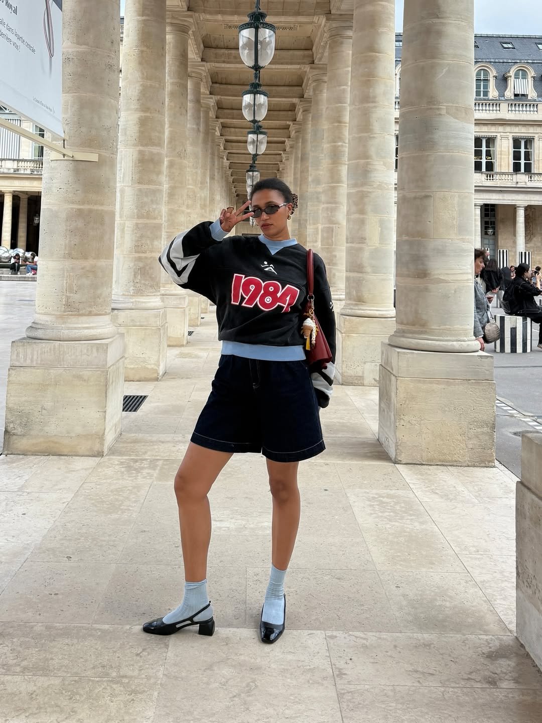 Pessoa vestida casualmente com camisa escura e sapatos de salto da moda, posando em um corredor de colunas elegantes.