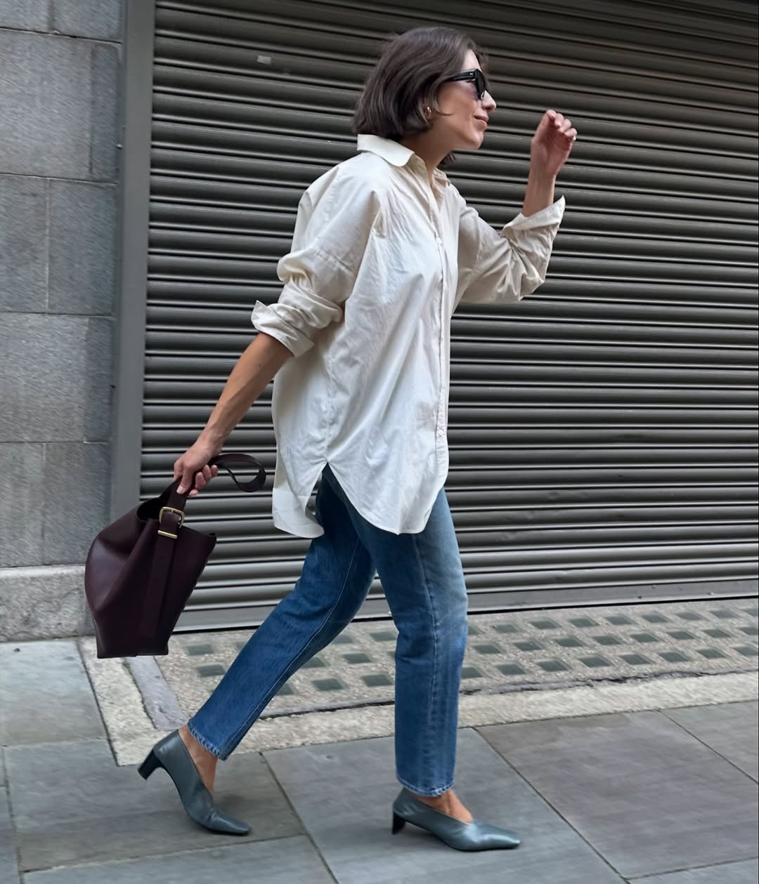 Mulher caminhando, usando sapatos de salto baixo, jeans e camisa larga, segurando uma bolsa marrom.
