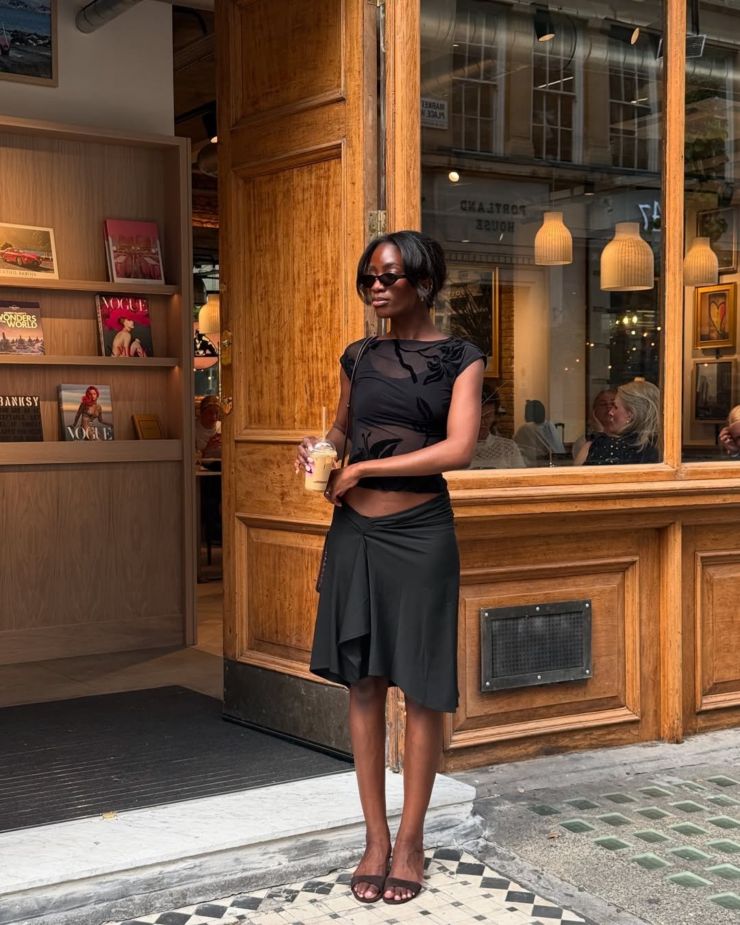 Mulher elegante com sapato de salto baixo e vestido preto segura bebida diante de loja charmosa.