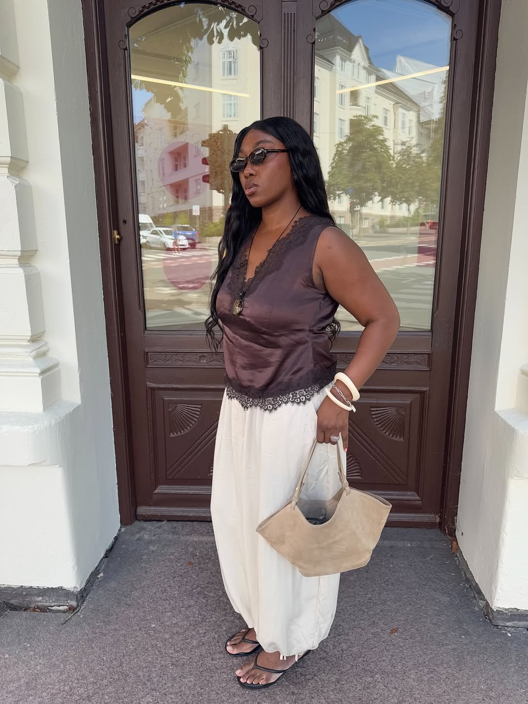 Mulher de blusa marrom e calça clara segura bolsa bege, usando sapato de salto baixo em frente a porta de madeira.