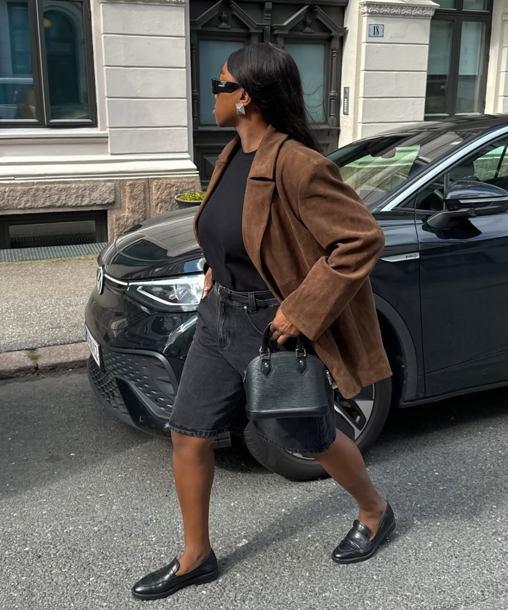 Mulher com jaqueta marrom e bermuda preta caminha na rua, carregando bolsa pequena preta. Ela usa sapatos estilosos.