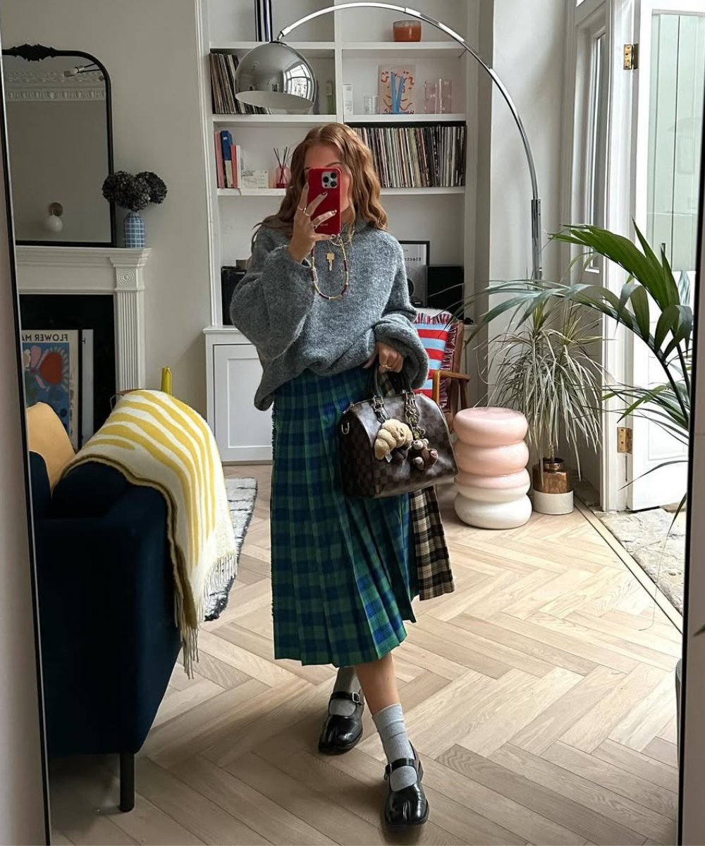 Usuária vestindo suéter cinza e saia xadrez azul e verde, com bolsa grande. Sapatos pretos completam o estilo.