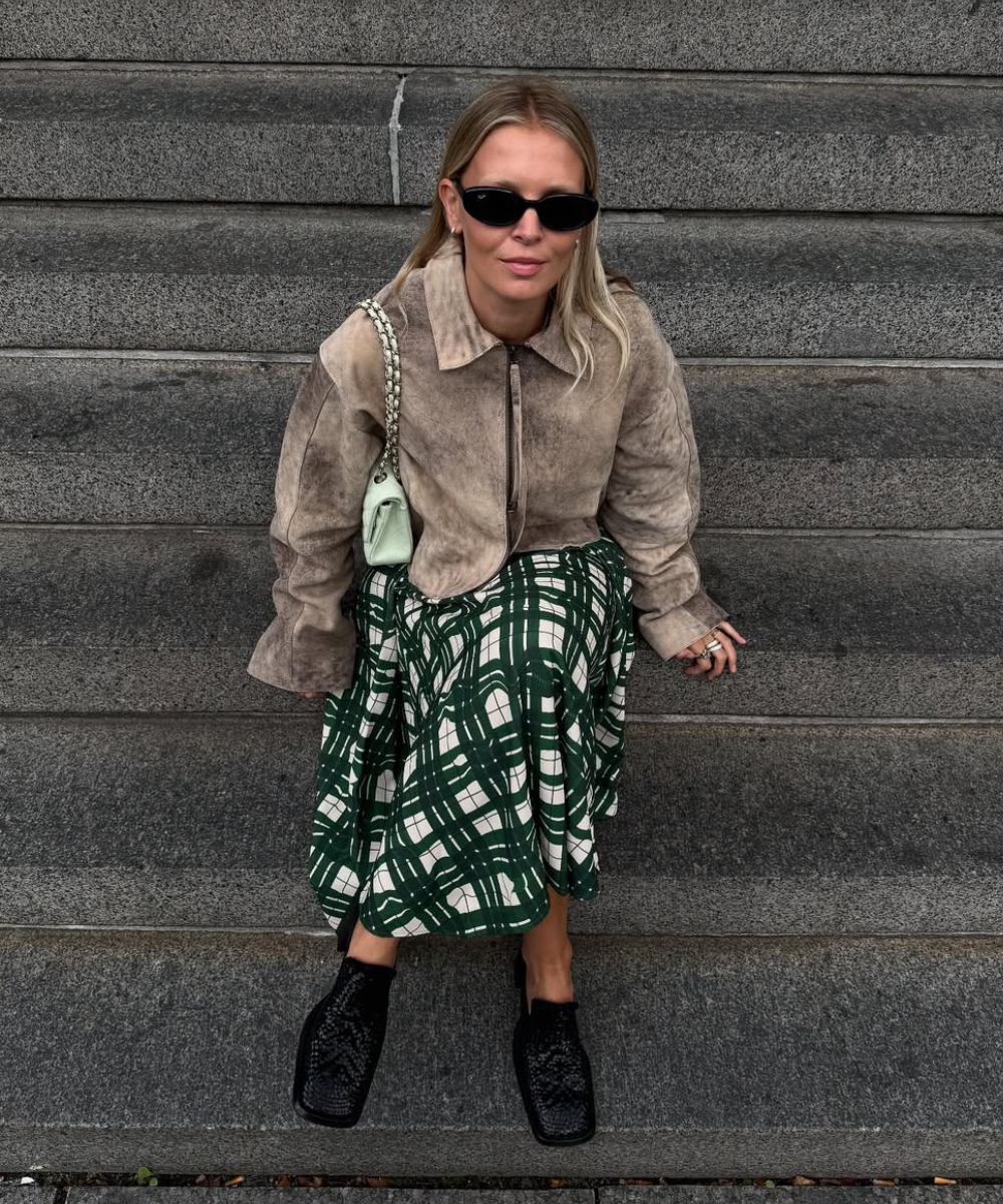 Pessoa sentada em escadas usando um casaco marrom, saia verde xadrez e sapatos ortopédicos estilosos pretos.