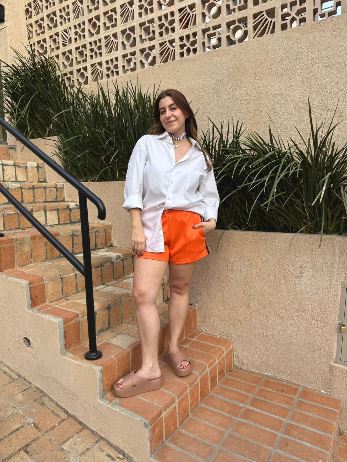 Mulher com camisa branca e shorts laranja, usando chinelos bege, uma das cores de sapato que vão bombar no verão de 2026, em escada de tijolos.
