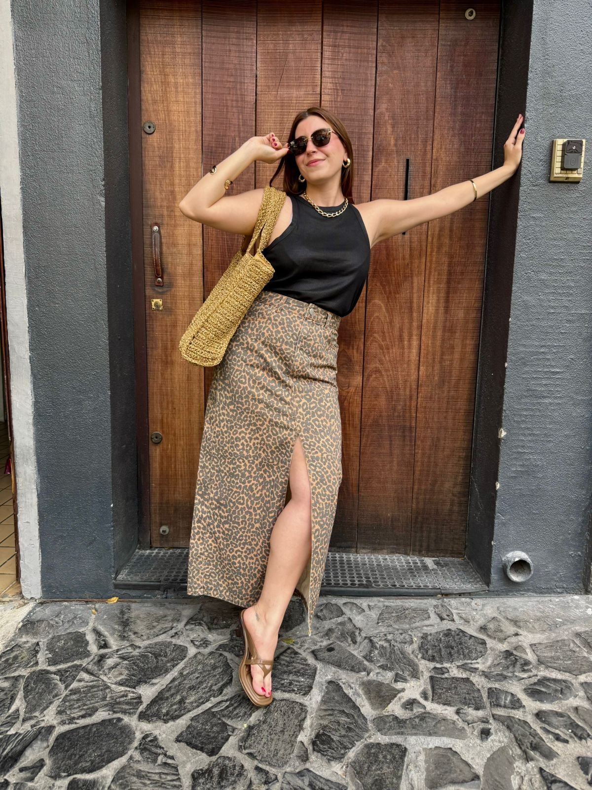 Pessoa com saia estampada e blusa preta, bolsa de palha, em frente à porta de madeira, e sapato marrom, uma das uma das cores de sapato que vão bombar no verão de 2026