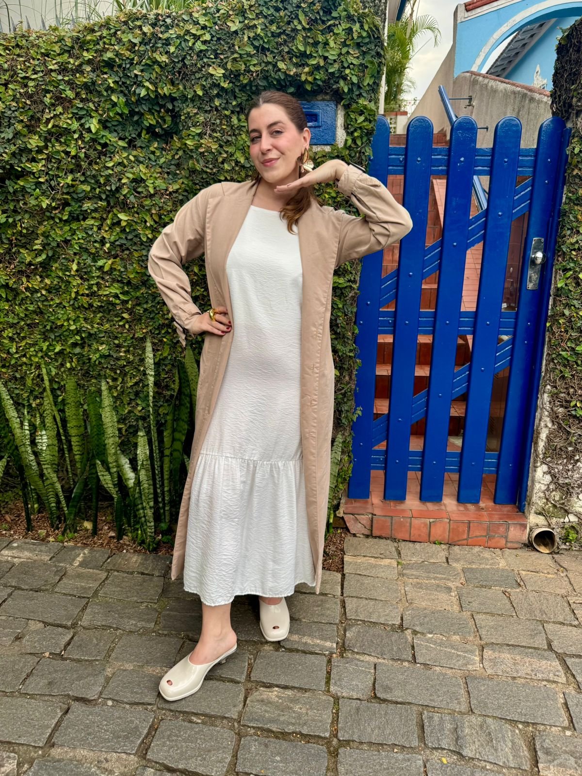 Mulher sorrindo em frente a um portão azul, usando vestido e cardigan com calçados claros, uma das cores de sapato que vão bombar no verão de 2026, destacando o look casual.