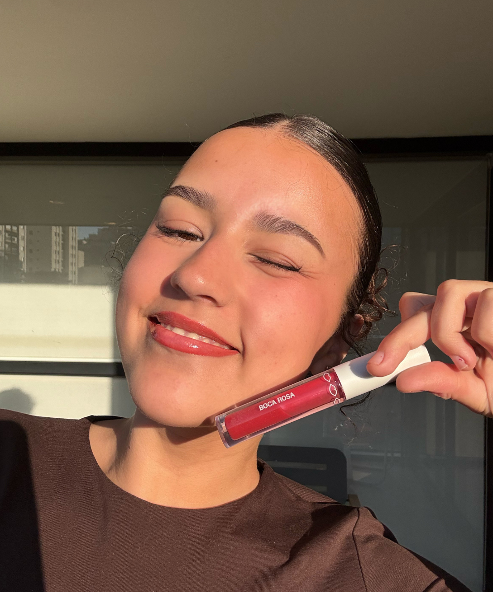 Sarah Fernandes sorrindo à luz do sol, segurando um batom líquido vermelho Hidra Lábios da Boca Rosa.