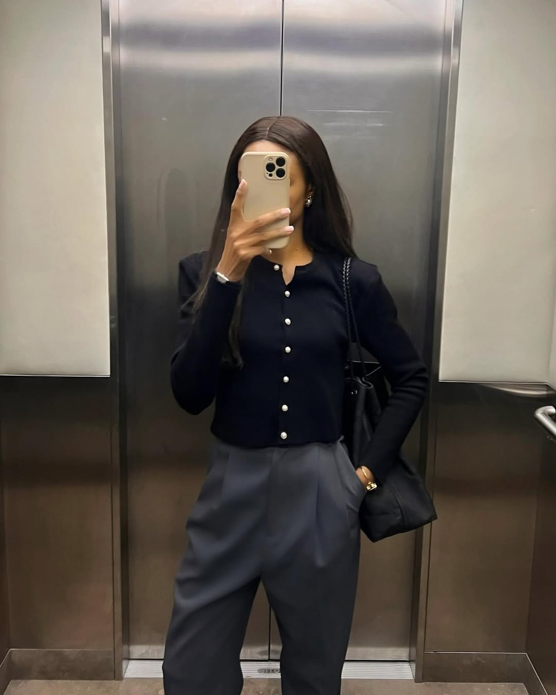 Pessoa usando traje formal e tirando selfie no espelho de um elevador, segurando bolsa preta.