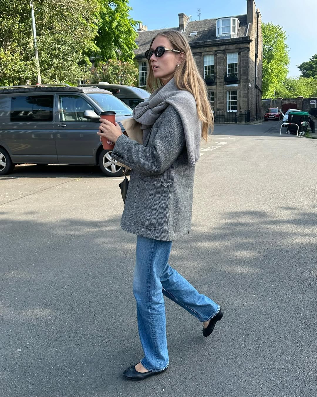 Uma mulher com casaco cinza e jeans segura café na rua.