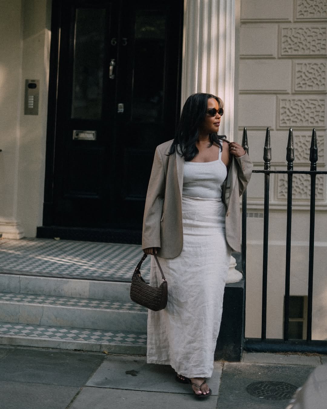 Mulher usando vestido longo branco, blazer bege e bolsa marrom, encostada em grade preta, em frente a porta escura.