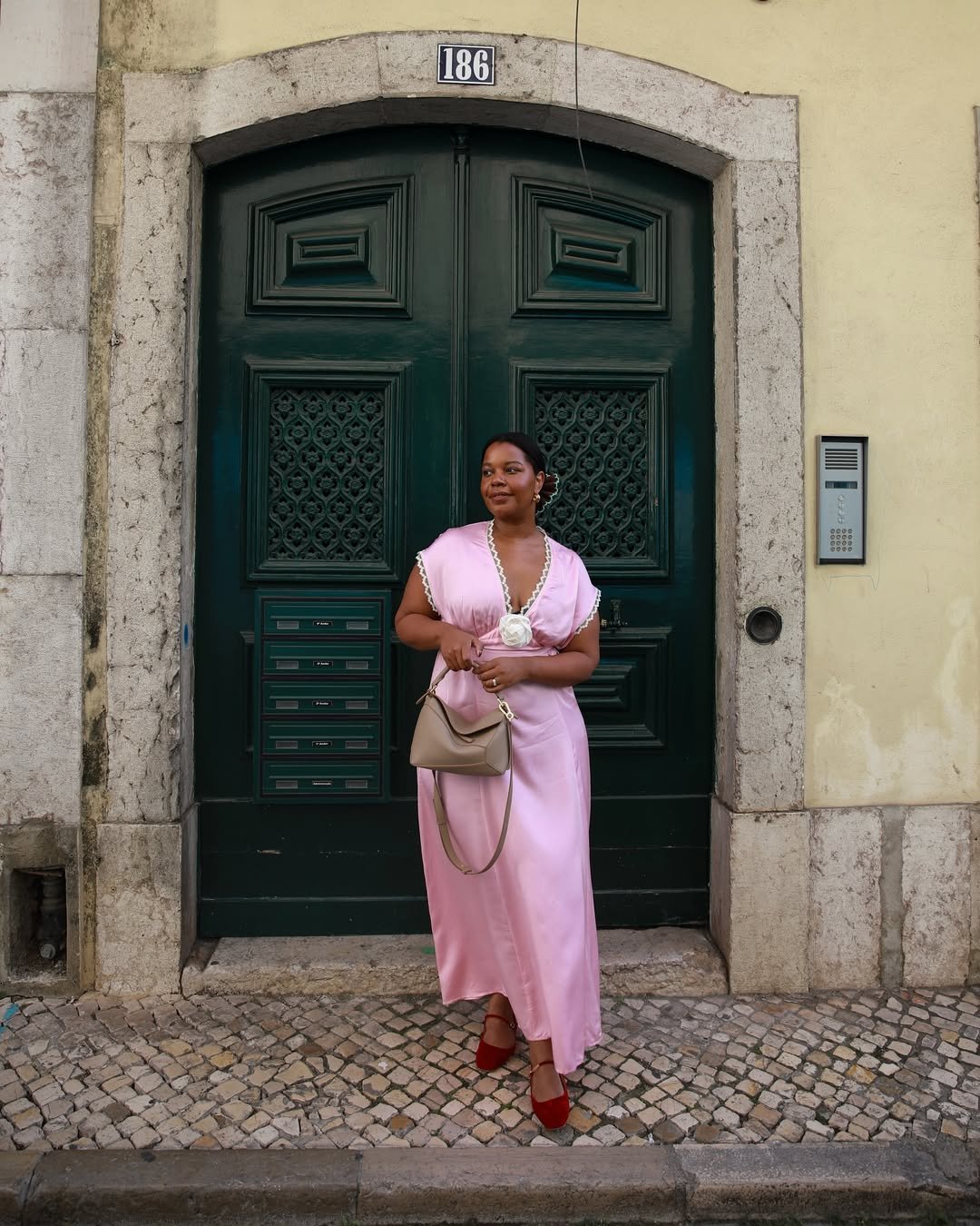 Mulher com vestido longo rosa, em pé diante de uma porta verde escura, segurando uma bolsa bege.