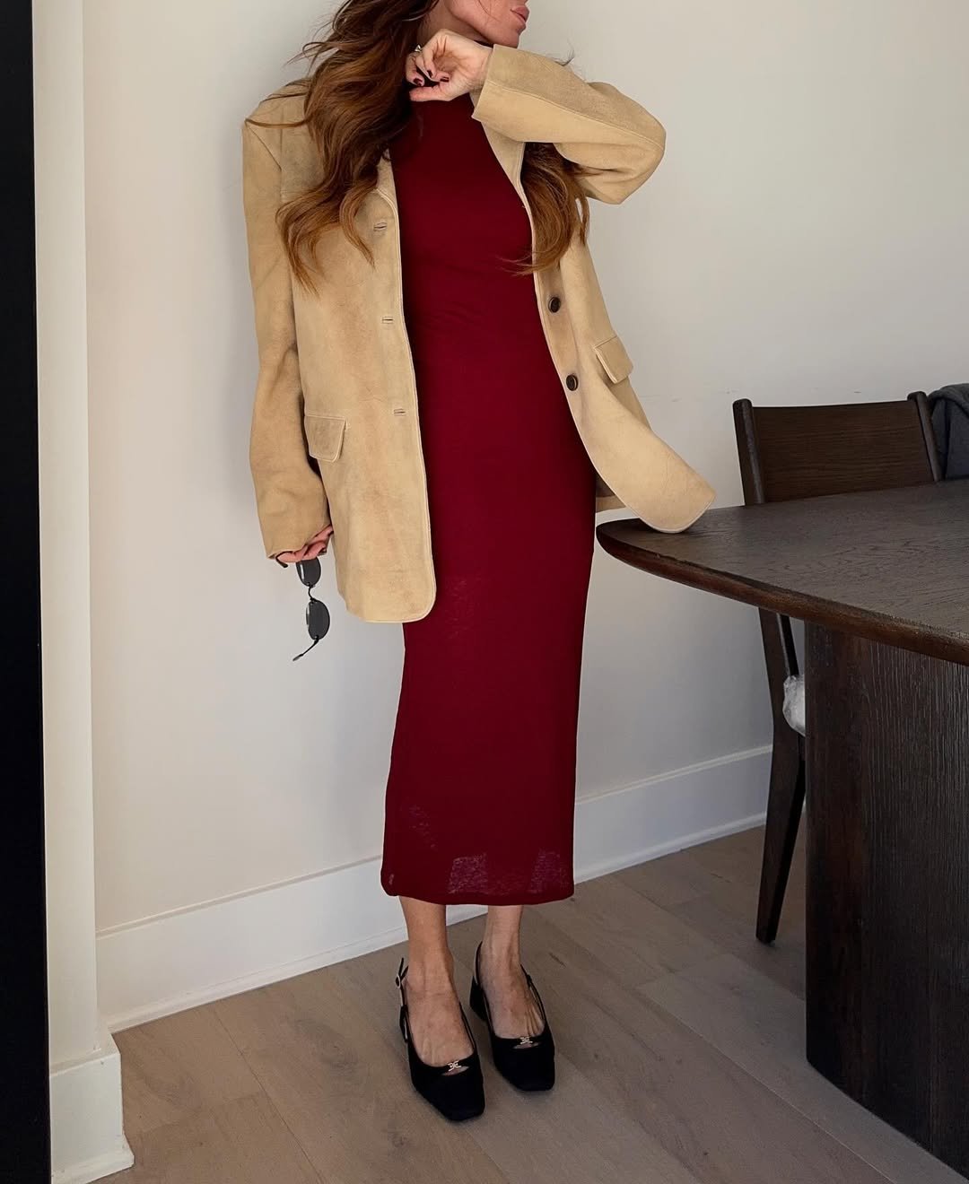 Mulher com vestido vermelho e blazer bege, posando ao lado de uma mesa, segurando óculos.